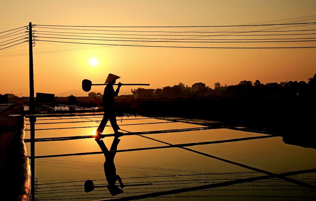 Golden sunset reflected across salt pans, silhouettes of workers in the distance.